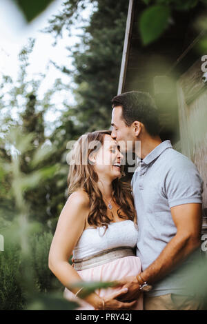 Il marito è baciare sua moglie incinta sulla sua fronte e sorride Foto Stock