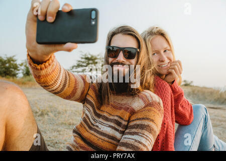 Un hipster paio di donna bionda e bruna uomo con capelli lunghi che indossa maglioni e rendendo selfie all'esterno. Foto Stock