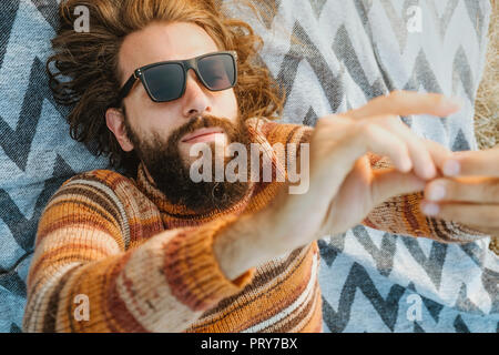 Un giovane adulto beardy uomo con capelli lunghi in un maglione e occhiali da sole agghiacciante sulla coperta all'aperto di sunrise Foto Stock