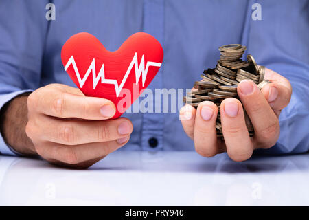 Close-up di una mano di un uomo azienda cuore rosso con la frequenza del polso e monete d'Oro Foto Stock