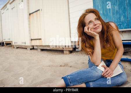 Redheaded donna seduta nella parte anteriore della cabina in spiaggia, la lettura di un libro Foto Stock