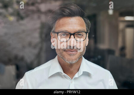 Ritratto di uomo con la stoppia dietro il vetro di finestra di indossare una camicia bianca e occhiali Foto Stock