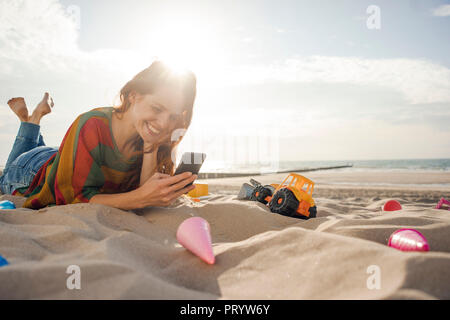 Redheaded donna sdraiata sulla spiaggia con giocattoli, utilizza lo smartphone Foto Stock