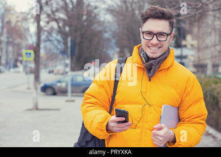 Giovane uomo bello nel rivestimento giallo tenendo lo smartphone e tablet camminando sulla strada e sorridente in telecamera Foto Stock