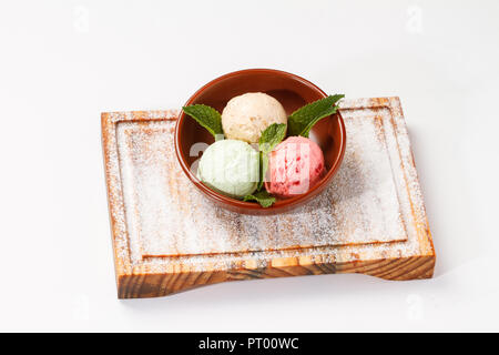 Gelato alla crema con le foglie di menta servito in una ciotola sulla parte superiore di una tavola di legno su sfondo bianco. 3 palline; lampone, pistacchio e caramello Foto Stock