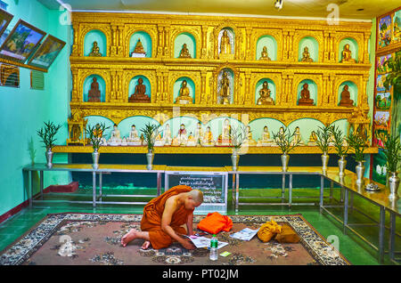 YANGON, MYANMAR - 27 febbraio 2018: il giovane monaco Bhikkhu legge il giornale in altare con donato immagini di Buddha nel tempio di Chaukhtatgyi, sul Fe Foto Stock