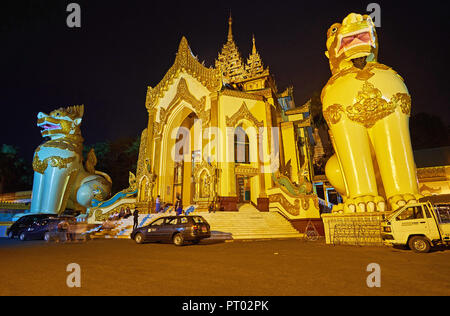 YANGON, MYANMAR - Febbraio 27, 2018: paesaggistico West Gate di Shwedagon pagoda con due giganti leogryphs (chinthe) guardie nella luminosa delle luci della sera, su F Foto Stock