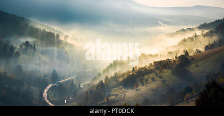 Panorama di paesaggi mozzafiato delle montagne di sunrise. incandescente nebbia salire dalla foresta su colline e rientrano nella valle. raggio di luce tracce di haze in s Foto Stock