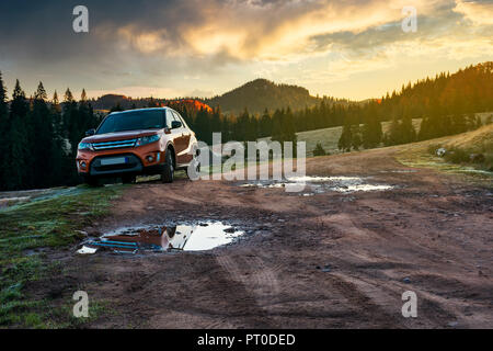 Orange suv parcheggiato sulla strada del paese vicino bosco in montagna a sunrise. bellissimo paesaggio autunnale. viaggia in Europa dal concetto di automobile Foto Stock