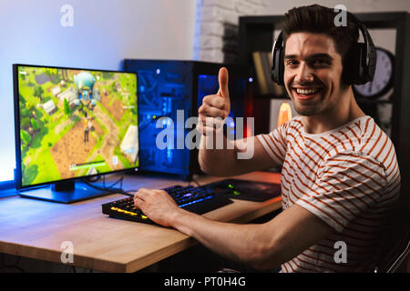 Ritratto di videogamer caucasica guy nelle cuffie per giocare con i videogiochi su computer e mostrando il pollice in alto Foto Stock