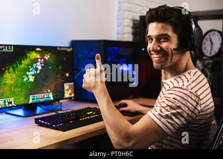 Ritratto di gioiosa gamer guy nelle cuffie per giocare con i videogiochi su computer e mostrando il pollice in alto Foto Stock