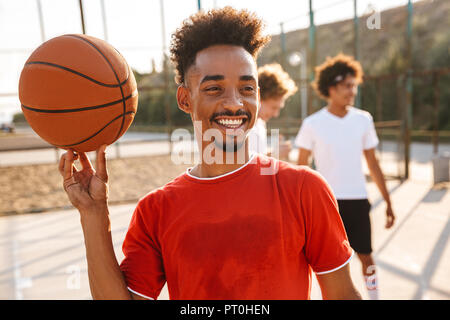 Ritratto di felice ragazzo africano sfera di filatura sul suo dito mentre giocare a basket presso il parco giochi all'aperto con il suo team Foto Stock