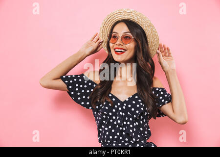 Lieta brunette donna nel cappello di paglia e occhiali da sole in posa mentre guarda lontano su sfondo rosa Foto Stock