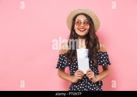 Incuriosito lieta brunette donna nel cappello di paglia e occhiali da sole azienda passaporto con biglietti mentre guarda lontano su sfondo rosa Foto Stock