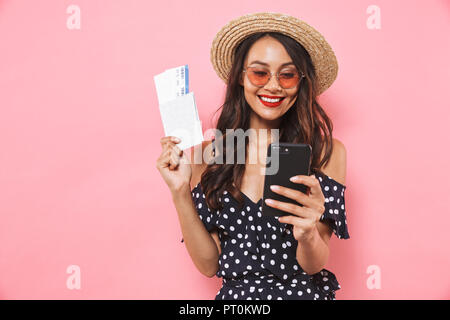 Lieta brunette donna nel cappello di paglia e occhiali da sole utilizza lo smartphone mentre si tiene il passaporto con i biglietti su sfondo rosa Foto Stock