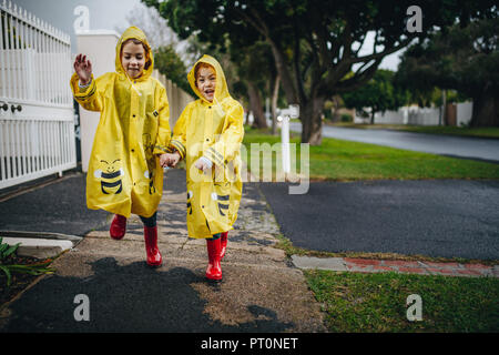 Due happy funny ragazze indossando un impermeabile giallo e rosso stivali di gomma. Gemelle alle passeggiate all'aperto in rivestimenti impermeabili. Foto Stock