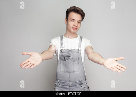 Ritratto di felice bruna giovane uomo in stile casual con t-shirt bianco jeans e tute da lavoro in piedi e guardando la telecamera con i bracci sollevati e sorridente Foto Stock