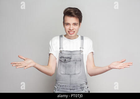 Non so. Ritratto di dubbia bruna giovane uomo in stile casual con t-shirt bianco jeans e tute da lavoro in piedi e guardando la fotocamera e confondere Foto Stock