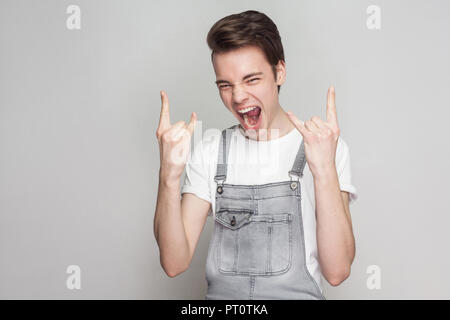 Stupiti crazy bruna giovane uomo in stile casual con t-shirt jeans e tute da lavoro in piedi e guardando la telecamera con gesto di roccia le mani e urlando. Foto Stock