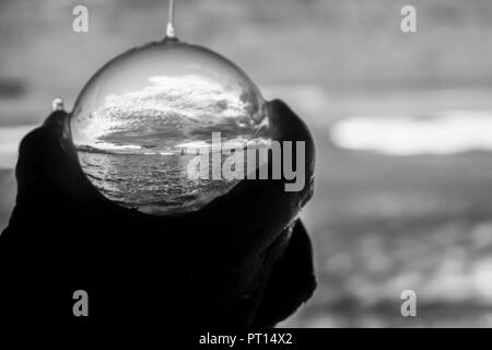 Argento e grigio sole e nuvole seascape con piccola barca sull orizzonte catturato in astratto Palla di vetro tenuto in mano con gocce d'acqua Foto Stock