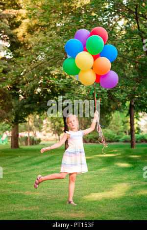Felice bambina gioca con grande grappolo di elio riempito palloncini colorati nel parco Foto Stock