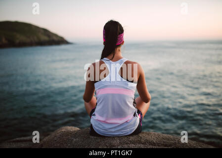 Attività sportive donna seduta sulle rocce, di fronte al mare di sera, vista posteriore Foto Stock