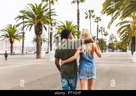 Spagna, Barcellona, vista posteriore del multiculturale di coppia giovane camminando a braccetto sul lungomare Foto Stock