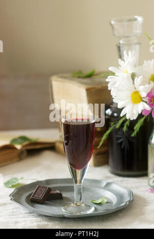 La crema de Cassis casalingo liquore di ribes nero in piccoli bicchieri, libri e fiori. Stile rustico. Foto Stock