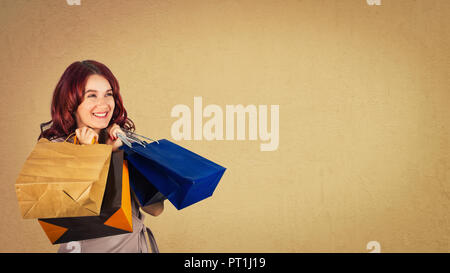 Excited donna giovane e bellissima azienda shopping bags sognante guardando lontano isolate su sfondo marrone con copia spazio. Foto Stock