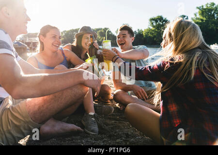 Gruppo di happy amici seduti in riva al fiume e la tostatura con bevande Foto Stock