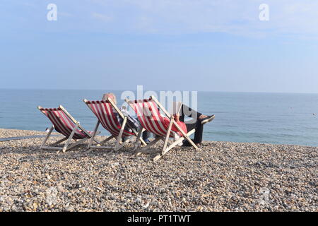 Matura in seduta a strisce rosse e bianche sedie a sdraio sulla spiaggia con ciottoli a birra durante estate indiana caldo sole di ottobre sul South Devon Coast, England, Regno Unito Foto Stock
