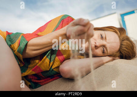 Redheaded donna sdraiata sulla spiaggia con sabbia passa attraverso la sua mano Foto Stock