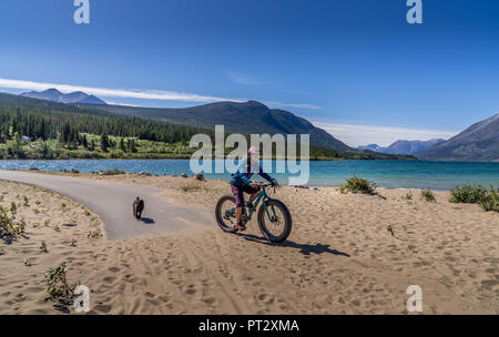 Una donna in bicicletta e a piedi il cane a Benet Lago e spiaggia di Carcross, Yukon Canada Foto Stock