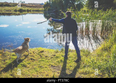 Un uomo con il Labrador retriever cane passeggiate sul lungolago in autunno. L'uomo treni il cane e getta il bastone in acqua Foto Stock