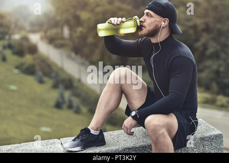 Muscolare atleta barbuto bere un acqua dopo una buona sessione di allenamento sul parco della città. Foto Stock