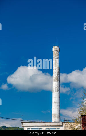 Dipinto di bianco di cemento senza fumo camino con asta di alleggerimento di un abbandono di fabbrica bulgara contro il cielo blu in una soleggiata giornata di primavera con soffici nuvole Foto Stock