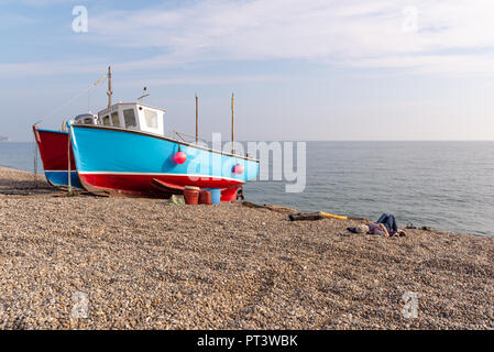 Donna sdraiata su una spiaggia di ciottoli accanto a due spiaggiata barche da pesca alla birra, Devon, Inghilterra, Regno Unito Foto Stock