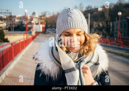 Ritratto di giovane donna che indossa down jacket e hat in piedi all'aperto accogliente avvolge se stessa fino a caldo sciarpa. Donna sorridente con gli occhi chiusi in piedi Foto Stock