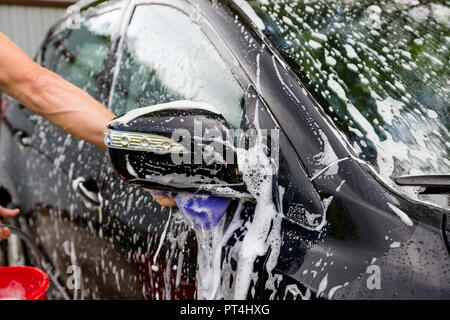 Pulizia automobile utilizzando acqua ad alta pressione. Lavaggio uomo la sua vettura sotto alta pressione acqua in service.lavoratore auto lavaggio.Protezione della concept car.mano maschio con spugna purpl il lavaggio auto.Il fuoco selettivo. Foto Stock