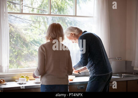 Vista posteriore a persone di mezza età amare giovane prepara la colazione insieme in cucina in piedi alla finestra grande, cura coppia marito aiutando senior moglie t Foto Stock