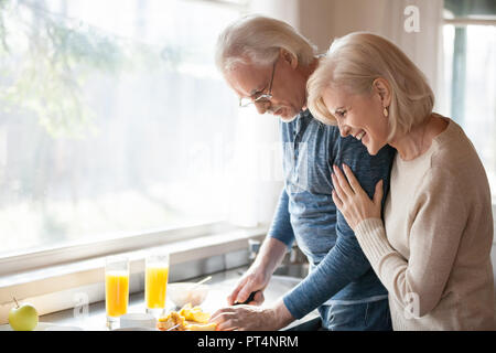 Caring senior marito preparare una sana fresca prima colazione al mattino per felice grato sorridente età moglie ridere abbracciando amare luomo in cucina, vecchio Foto Stock