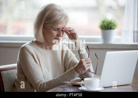 Sconvolto affaticato oberati di lavoro senior business maturo donna prendendo il largo bicchieri stanco del computer di lavoro, esausto di mezza età soffre dipendente da sfocata v Foto Stock