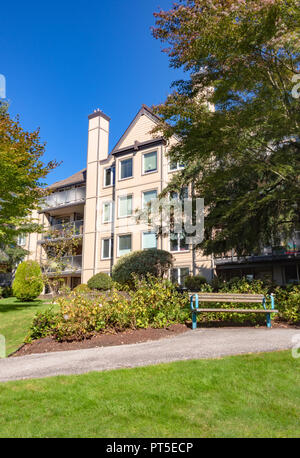 Basso edificio residenziale e panca in legno nella parte anteriore. Edificio di appartamenti e piccola area parco sulla giornata di sole Foto Stock