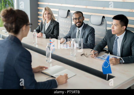 Fiducioso positivo multi-etnico politici in abiti formali seduti al tavolo da conferenza con le bandiere nazionali e la visualizzazione di documenti con grafici mentre discu Foto Stock