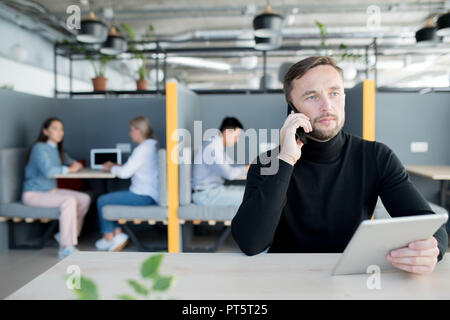Grave pensieroso successo manager maschio in maglione nero seduta a tavola e tenere compressa mentre parla al telefono in ufficio Foto Stock