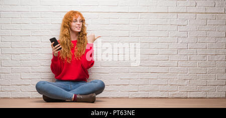 Giovani redhead donna seduta su un muro di mattoni a parlare al telefono di puntamento e mostra con il pollice fino a che il lato con buon viso sorridente Foto Stock