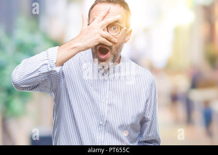 La mezza età annoso senior uomo che indossa gli occhiali su sfondo isolato peeking in stato di shock che copre il viso e gli occhi con la mano, guardando attraverso le dita con e Foto Stock