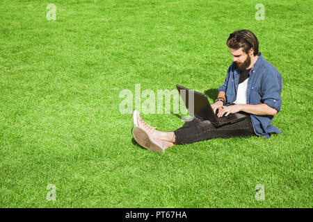 Fiducioso barbuto giovane uomo in jeans shirt indossando occhiali da sole seduti all'aperto sull'erba verde e lavorando diligentemente Foto Stock