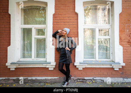 Fashion lifestyle Ritratto di giovane donna alla moda vestito di mantello nero, blu berretto lavorato a maglia in posa, ridendo e sorridente nella città vecchia, autunno street Foto Stock