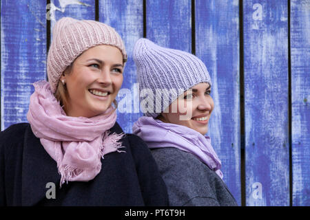 Ritratto di due bellissimi modelli giovani in blu e rosa cappelli a maglia in piedi vicino a parete blu su una giornata autunnale. Autunno Caldo foto.Donna sorridente e Foto Stock